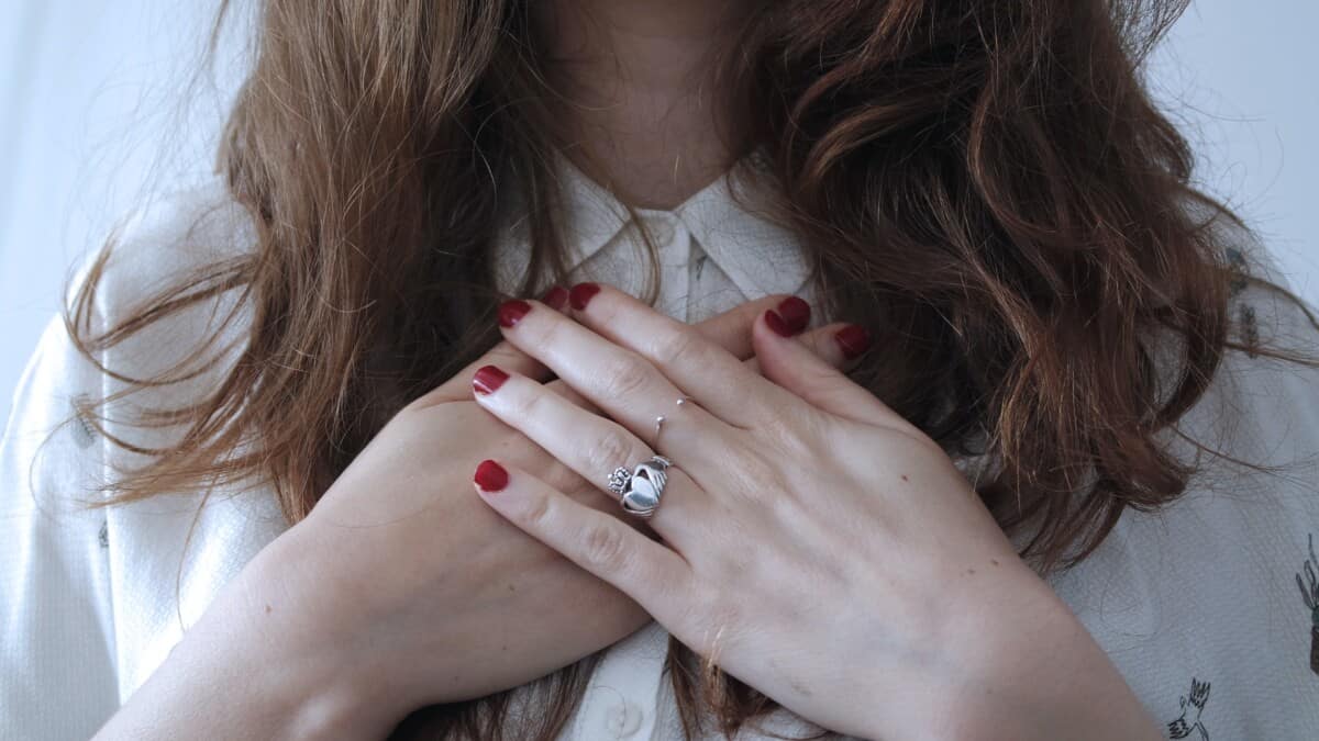 Een vrouw met lang bruin haar kruist haar handen over haar borst, toont rode nagellak en zilveren ringen, terwijl ze een wit overhemd draagt.
