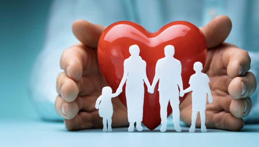 Een paar handen die een rood hart vasthouden achter witte papieren knipsels van een gezin met twee volwassenen en twee kinderen, symbool voor bescherming, zorg en gezondheid van het gezin.