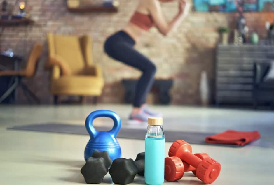 Een vrouw oefent binnenshuis op een yogamat, onscherp op de achtergrond. Op de voorgrond liggen een blauwe kettlebell, zwarte handgewichten, rode halters, een fles blauwe vloeistof en een opgerolde oranje handdoek.