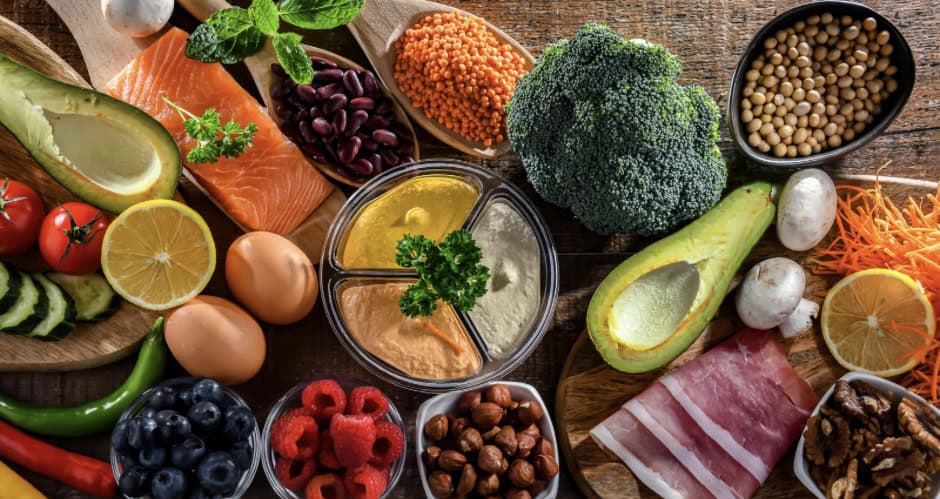 Een verscheidenheid aan gezond voedsel op een houten tafel, waaronder avocado, zalm, eieren, linzen, bonen, broccoli, noten, bessen, gesneden groenten, halve citroenen, ham en verschillende dipsauzen.
