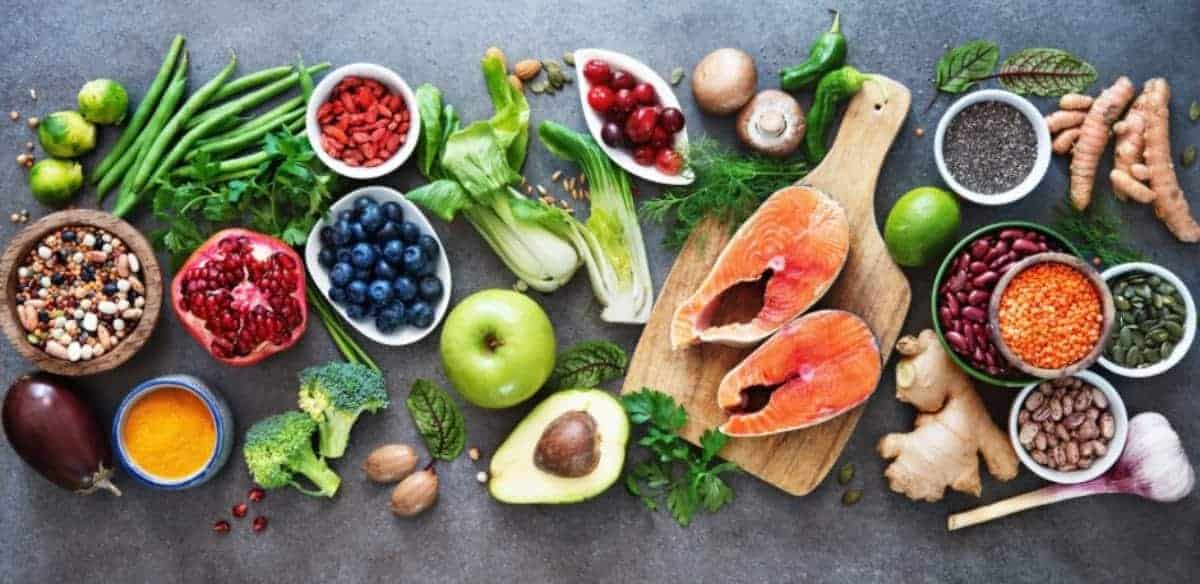Een verscheidenheid aan vers fruit, groenten, zaden, noten en twee rauwe zalmfilets op een grijs oppervlak, die gezonde, kleurrijke en voedzame voedingsopties laten zien.
