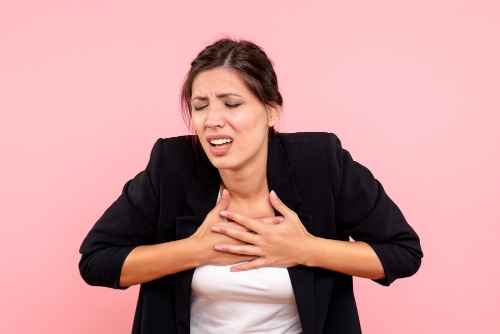 Een vrouw met donker haar in een zwarte blazer en wit shirt houdt met beide handen haar borst vast en lijkt pijn of ongemak te hebben, staande tegen een roze achtergrond.