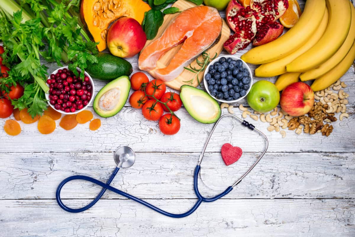 Een verscheidenheid aan gezonde voedingsmiddelen, waaronder fruit, groenten, noten en zalm, zijn gerangschikt op een rustieke witte houten tafel met een stethoscoop en een klein rood hart op de voorgrond.
