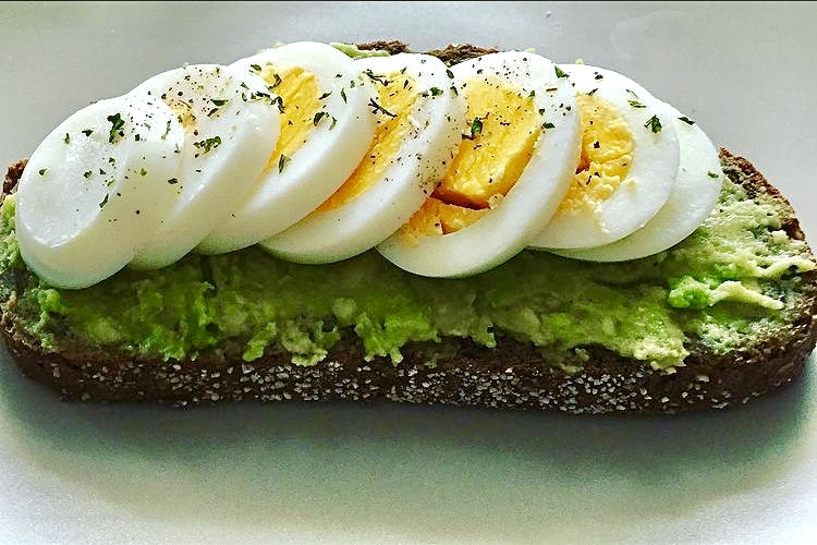 Een sneetje donker brood met daarop gepureerde avocado en verschillende plakjes hardgekookt ei, bestrooid met kruiden, op een wit bord.