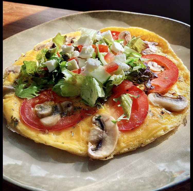 Een gouden omelet met daarop verse sla, komkommerblokjes, rode paprika, plakjes tomaat en plakjes champignon, geserveerd op een beige bord onder natuurlijk licht.