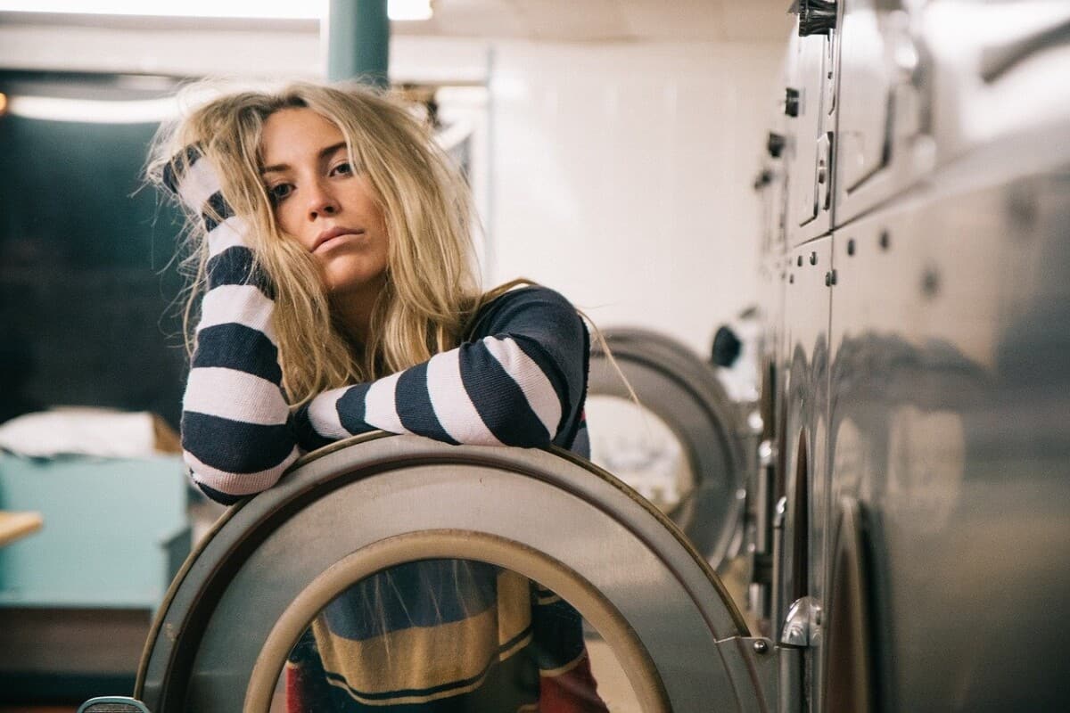 Een vrouw met lang blond haar en een gestreept shirt leunt met haar hoofd op haar hand terwijl ze haar arm laat rusten op een wasmachine in een wasserette, ze ziet er moe of verveeld uit.