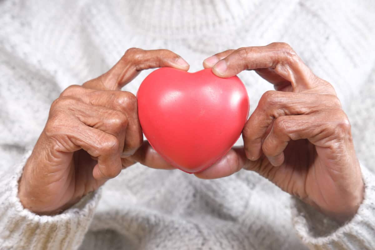 Een oudere persoon met een witte trui houdt een rood hartvormig voorwerp voorzichtig met beide handen op borsthoogte.