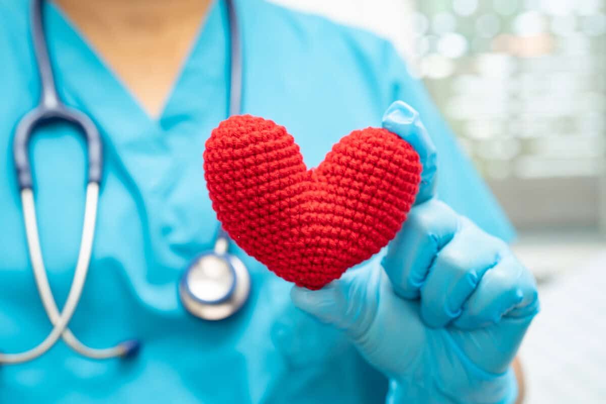 Een medewerker in de gezondheidszorg draagt blauwe scrubs en handschoenen en houdt een rood, gebreid hart in één hand. Om hun nek hangt een stethoscoop, symbool voor zorg en gezondheid van het hart.