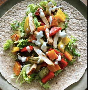 Een volkoren tortilla belegd met gegrilde kipreepjes, gemengde kleurrijke paprika's, sla en een drupje romige witte dressing, geserveerd op een groen bord.