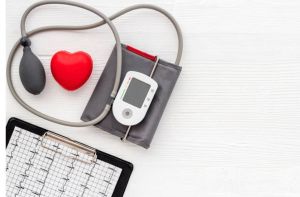 Een bloeddrukmeter met manchet, een rood hartvormig voorwerp en een ECG-kaart op een klembord worden op een wit houten oppervlak geplaatst.