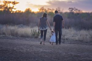Een man en vrouw lopen hand in hand met een klein kind in een witte jurk langs een zandpad buiten bij zonsondergang, met bomen en grassen op de achtergrond.