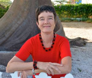 Een vrouw met kort bruin haar en een felrood topje, bijpassende kralenketting en armband zit buiten voor een grote boomstam en lacht naar de camera.