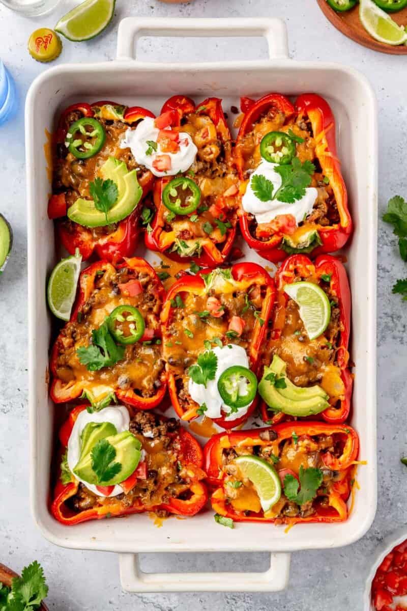 Een ovenschaal gevuld met gevulde rode paprika's met daarop gesmolten kaas, avocadoplakjes, jalapeños, zure room, partjes limoen, tomaten en verse koriander.