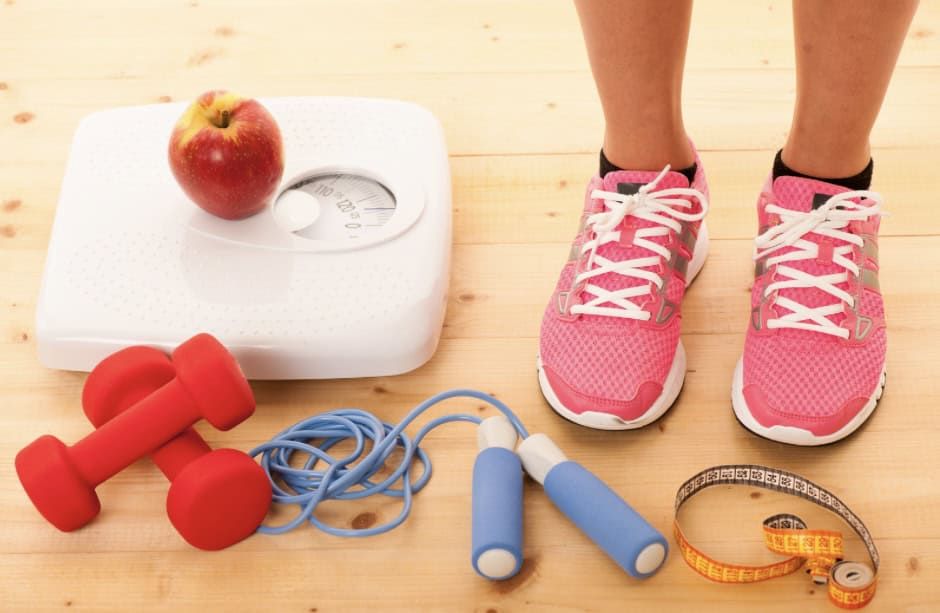 Persoon met roze gymschoenen staat naast een weegschaal met een appel erop, rode halters, een blauw springtouw en een geel meetlint op een lichte houten vloer.